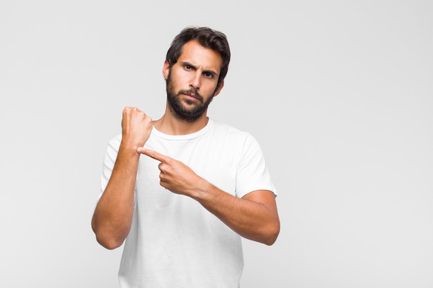 Joven guapo latino que parece impaciente y enojado, apuntando al reloj, pidiendo puntualidad, quiere llegar a tiempo