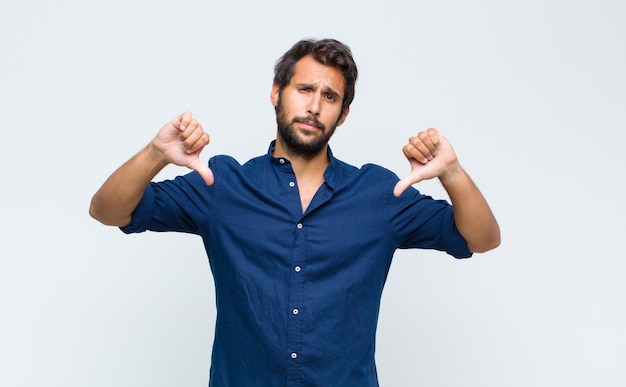 Joven guapo latino que se siente enfadado, enojado, molesto, decepcionado o disgustado, mostrando los pulgares hacia abajo con una mirada seria