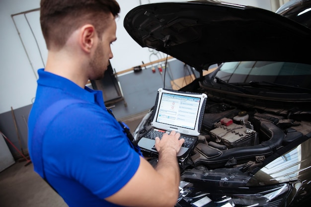 Joven y guapo mecánico de automóviles confiado con una computadora portátil en la mano realiza diagnósticos por computadora para detectar errores en los sistemas del automóvil