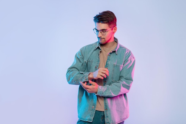 Un joven guapo a la moda con peinado y gafas elegantes usa una camisa de mezclilla de moda en el estudio con luces creativas rosas y azules