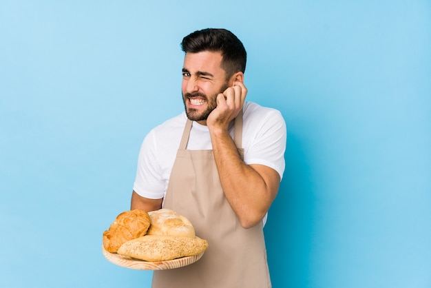 Joven guapo panadero aislado cubriendo las orejas con las manos.