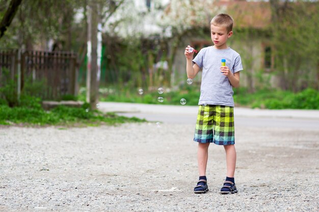 Joven guapo rubio colegial en ropa casual con divertida expresión grave sopla bombillas de jabón transparente de pie al aire libre en un cálido día de verano en la escena rural. Descuidado concepto de infancia feliz.