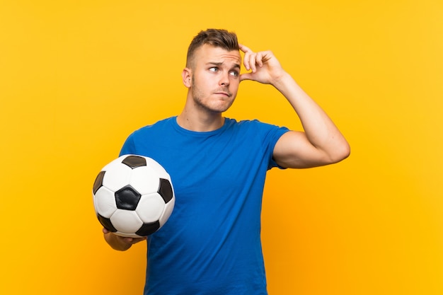 Joven guapo rubio sosteniendo un balón de fútbol con dudas y con expresión de la cara confusa