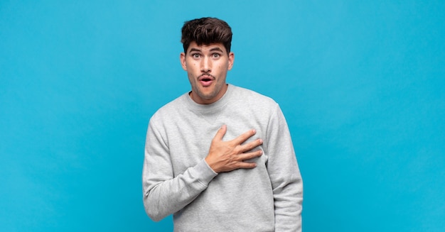 Joven guapo sintiéndose conmocionado y sorprendido, sonriendo, tomando la mano al corazón, feliz de ser el indicado o mostrando gratitud