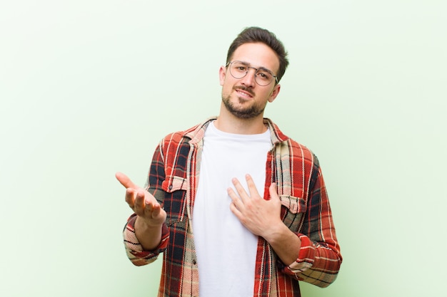 Joven guapo sintiéndose feliz y enamorado, sonriendo con una mano al lado del corazón y la otra estirada al frente