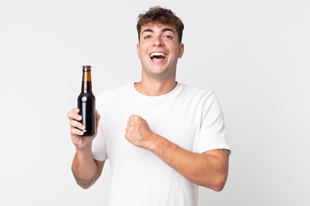 Joven guapo sintiéndose feliz y enfrentando un desafío o celebrando y sosteniendo una botella de cerveza