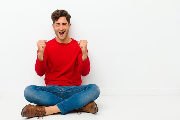 Joven guapo sintiéndose feliz, sorprendido y orgulloso, gritando y celebrando el éxito con una gran sonrisa sentada en el suelo