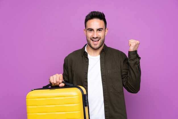 Joven guapo sobre pared púrpura aislada en vacaciones con maleta de viaje y un sombrero