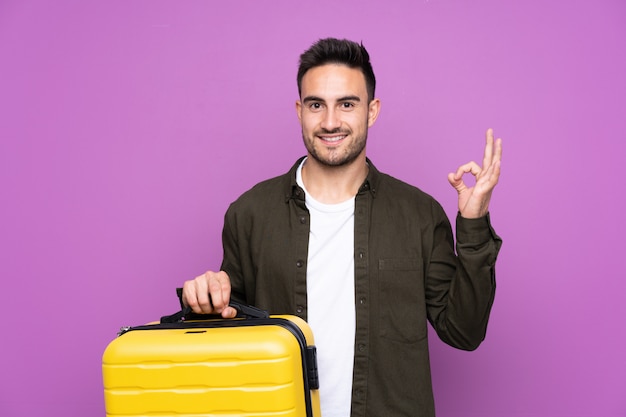 Joven guapo sobre púrpura en vacaciones con maleta de viaje y un sombrero