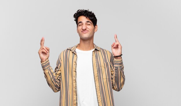 Joven guapo sonriendo y cruzando ambos dedos con ansiedad, sintiéndose preocupado y deseando o esperando buena suerte