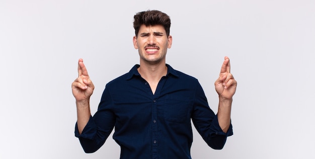 Joven guapo sonriendo y cruzando ambos dedos con ansiedad, sintiéndose preocupado y deseando o esperando buena suerte