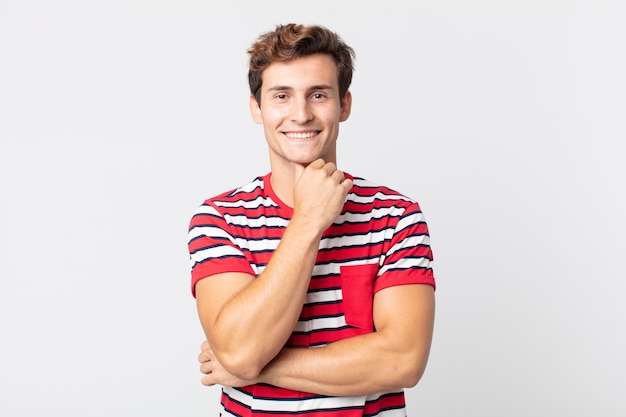 Joven guapo sonriendo, disfrutando de la vida, sintiéndose feliz, amigable, satisfecho y despreocupado con la mano en la barbilla