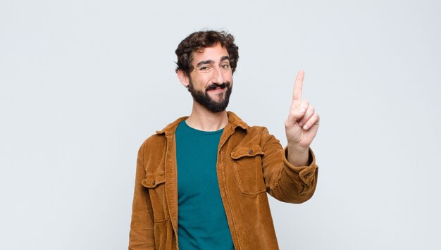 Joven guapo sonriendo y mirando amistosamente, mostrando el número uno o el primero con la mano hacia adelante, contando hacia atrás contra la pared plana