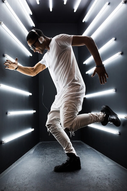 Joven guapo sonriente hipster negro en traje blanco escuchando música en auriculares y bailando al estilo hip hop en discoteca, divirtiéndose