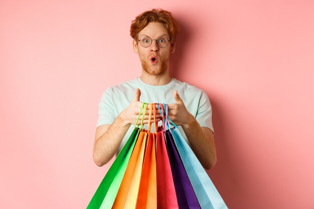 Joven guapo sorprendido con descuentos especiales, comprando regalos, sosteniendo bolsas de compras y de pie sobre fondo rosa.