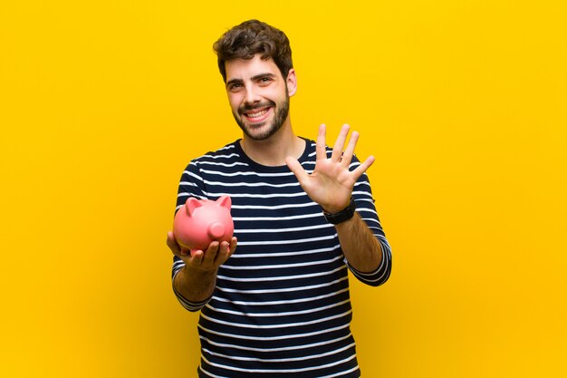 Joven guapo sosteniendo una alcancía contra el fondo naranja