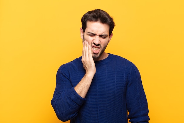 Joven guapo sosteniendo la mejilla y sufriendo dolor de muelas, sintiéndose enfermo, miserable e infeliz, buscando un dentista