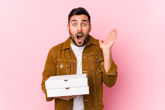 Joven guapo sosteniendo pizzas aislado sorprendido y conmocionado.