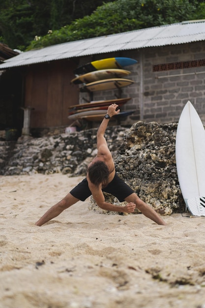 Un joven y guapo surfista en la orilla del océano está calentando antes de surfear. ejercicios antes del deporte, estiramientos antes del surf.