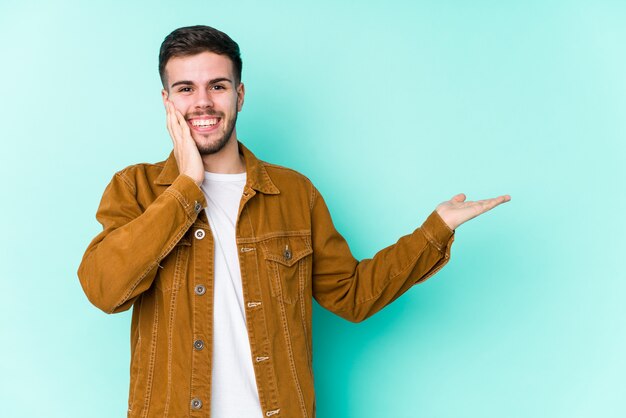 Joven guapo tiene espacio de copia en una palma, mantenga la mano sobre la mejilla Sorprendido y encantado.