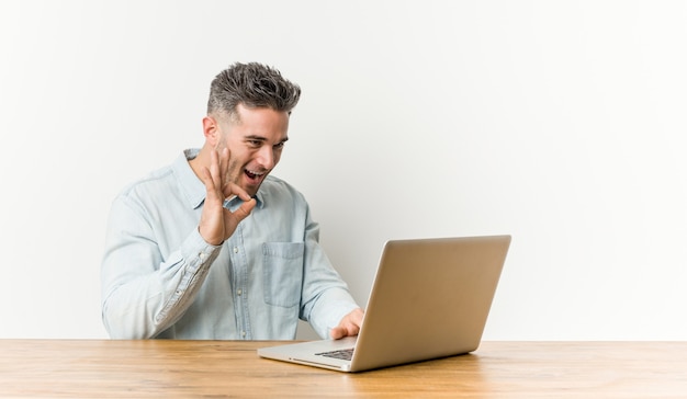 Joven guapo trabajando con su computadora portátil guiña un ojo y tiene un gesto bien con la mano.