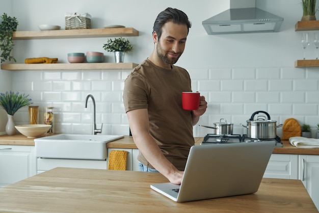 Un joven guapo usando una computadora portátil y disfrutando del café mientras está de pie en la isla de la cocina