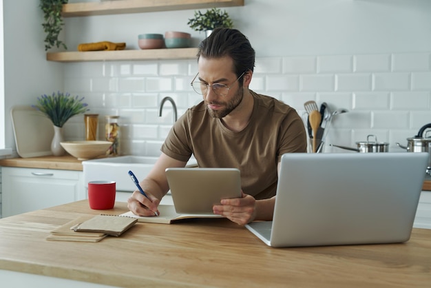 Un joven guapo usando tecnologías mientras trabaja en la isla de la cocina en casa