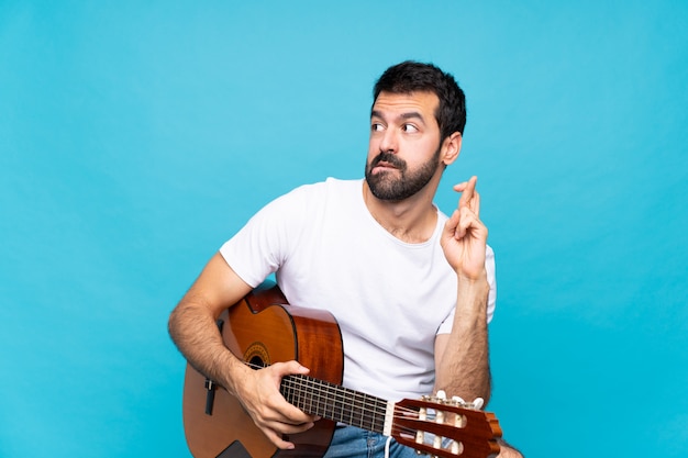 Joven con guitarra sobre azul aislado con dedos cruzando y deseando lo mejor