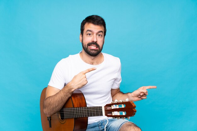 Joven con guitarra sobre fondo azul aislado asustado y apuntando hacia el lado