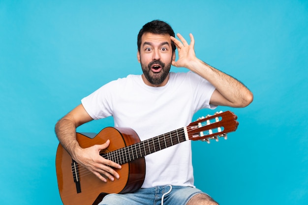 Joven con guitarra sobre pared azul aislada con expresión de sorpresa