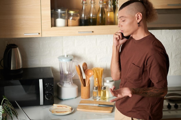 Joven hablando por teléfono con un colega cuando prepara el desayuno en la cocina