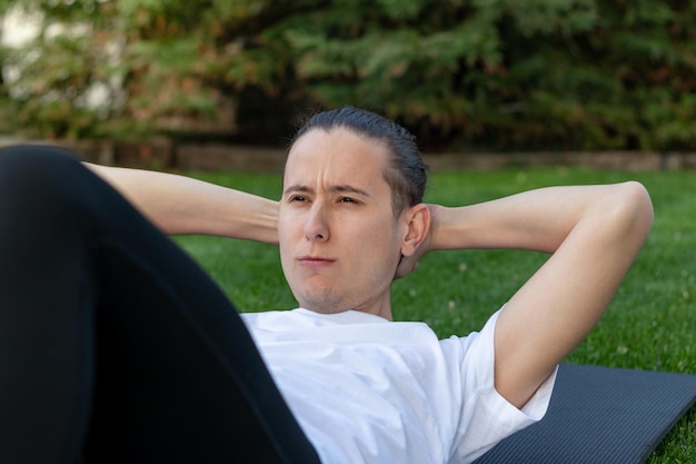 Joven haciendo abdominales con cara de sufrimiento y esfuerzo mientras está en el jardín al aire libre