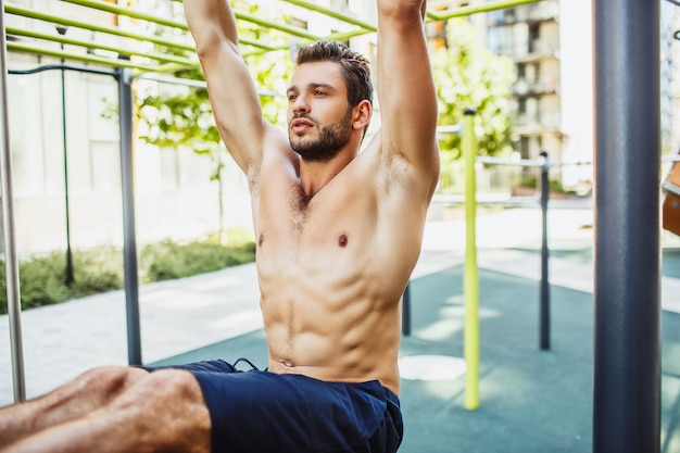 Joven haciendo ejercicio afuera. Cerrar y cortar la vista de un tipo musculoso fuerte, poderoso y bien construido que cuelga de la barra horizontal