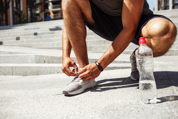 Joven haciendo ejercicio afuera. Corte la vista baja de los cordones de los zapatos o zapatillas de deporte del hombre. Botella de agua casi vacía además sobre asfalto.