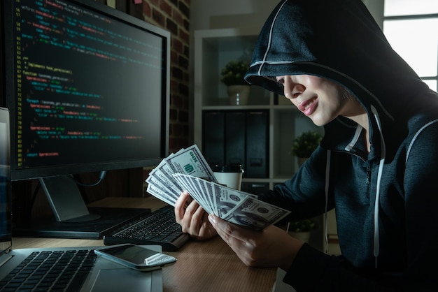 joven hacker con dinero en efectivo en las manos. sonriendo con saña y contando la cantidad de dinero.