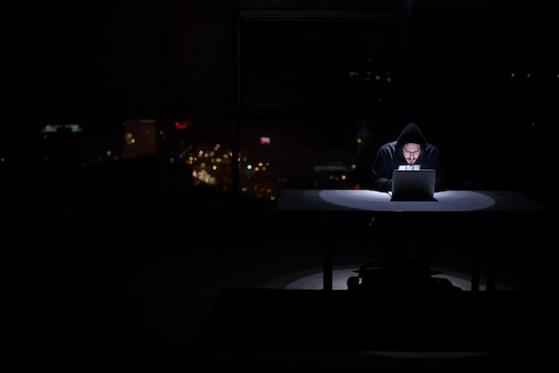 Joven hacker talentoso que usa una computadora portátil mientras trabaja en una oficina oscura con las luces de la gran ciudad en el fondo por la noche