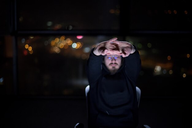 Joven hacker talentoso que usa una computadora portátil mientras trabaja en una oficina oscura con las luces de la gran ciudad en el fondo por la noche