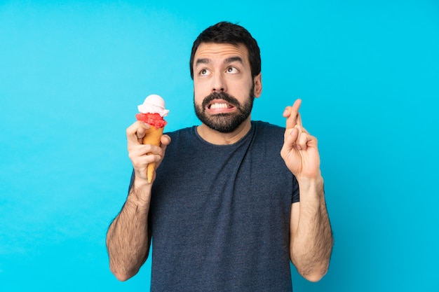 Joven con un helado de cucurucho sobre pared azul aislado con dedos cruzando y deseando lo mejor