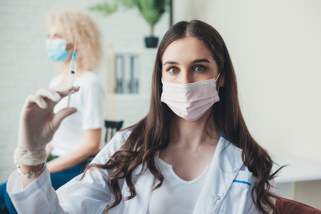 Joven hermana médica lleva una máscara y sostiene una vacuna mirando a la cámara