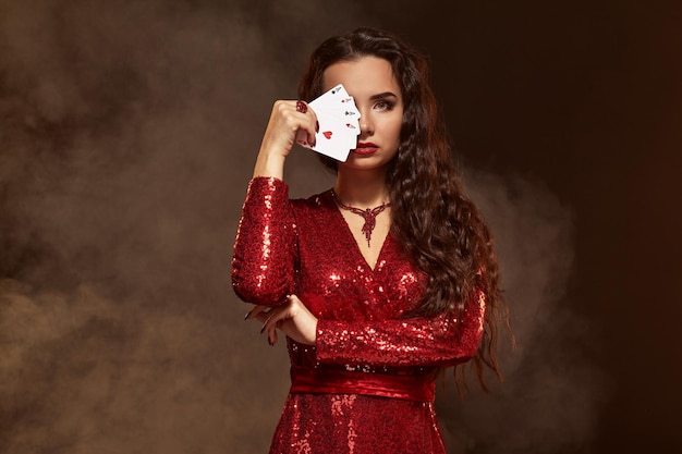 Joven hermosa afortunada morena con un vestido de noche rojo muestra sus cartas un cuatro de ases escondiendo su rostro ...