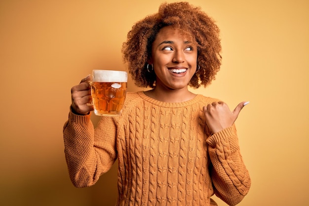 Joven hermosa afroamericana afro con cabello rizado bebiendo jarra de cerveza señalando y mostrando con el pulgar hacia un lado con cara feliz sonriendo