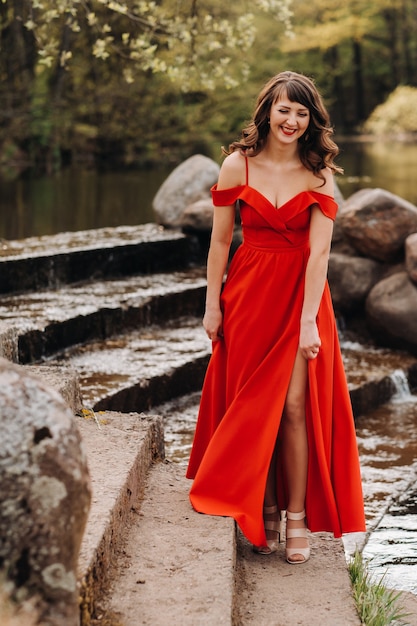 Una joven hermosa con cabello largo castaño, con un vestido largo rojo, baja las escaleras cerca del lago,