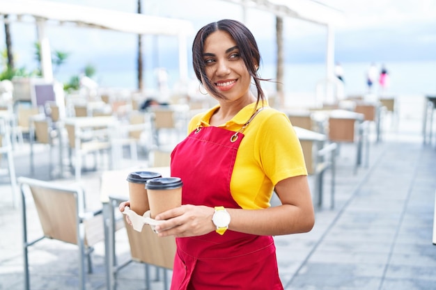 Joven hermosa camarera árabe sonriendo confiada sosteniendo café para llevar en la terraza de la cafetería