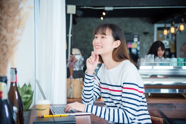 Joven hermosa chica asiática sonriendo mientras trabajaba con la computadora portátil en la cafetería