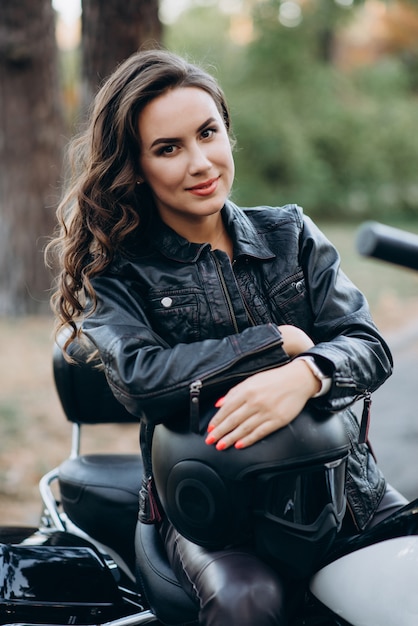 Joven hermosa chica biker en una chaqueta de cuero negro en una motocicleta con un casco en sus manos. El concepto de velocidad y libertad.