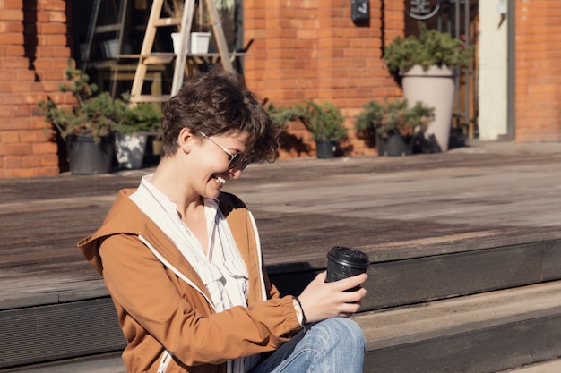 Joven hermosa chica con un elegante corte de pelo se sienta en la calle y bebe café.