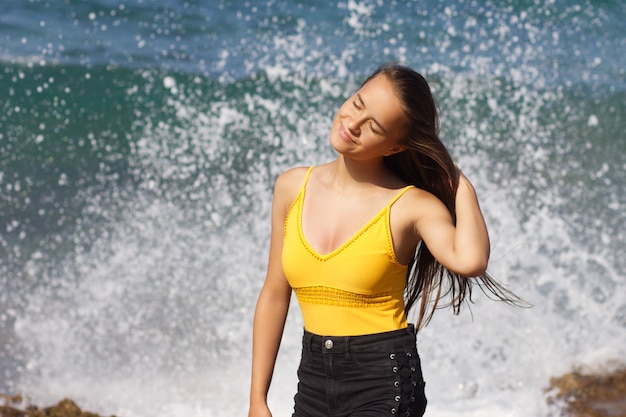 Joven hermosa chica europea de pelo oscuro en traje de baño amarillo sonriendo y de pie en el fondo del mar y salpicaduras en un día soleado