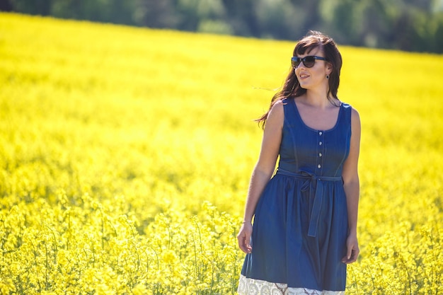 Joven hermosa chica feliz con un vestido azul con gafas de sol en un floreciente campo de colza en verano