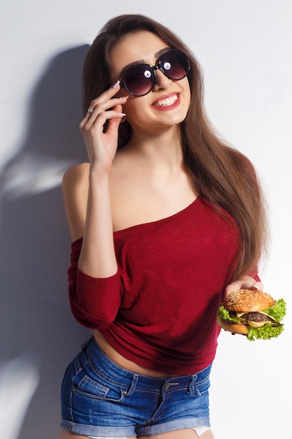 Joven hermosa chica con una hamburguesa en sus manos. Foto de estudio Modelo. Comida de comida rápida. Chica vestida con pantalones cortos y una camiseta, gafas de sol en la cara. El concepto de comida.