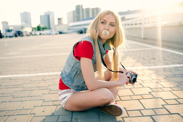 Foto joven hermosa chica hipster de moda sexy en pantalones cortos está descansando sentada en el suelo y sopla burbujas de chicle y tomando fotografías el concepto de estilo de vida